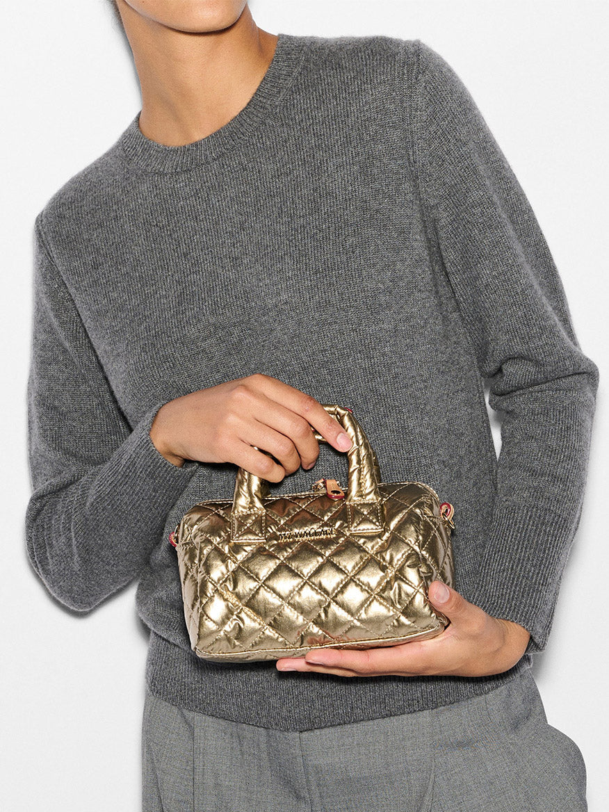 A person in a grey sweater holds a stylish MZ Wallace Mini Metro Satchel in Antique Gold Metallic Oxford, crafted from natural Italian leather.