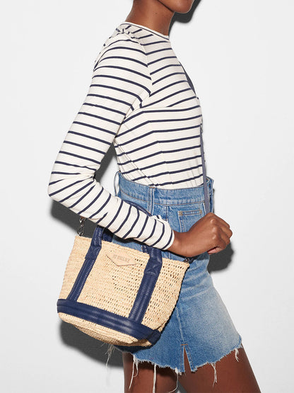 A person wearing a striped long-sleeve shirt and denim overalls carries a MZ Wallace Mini Raffia Tote in Raffia/Navy with padded nylon handles.