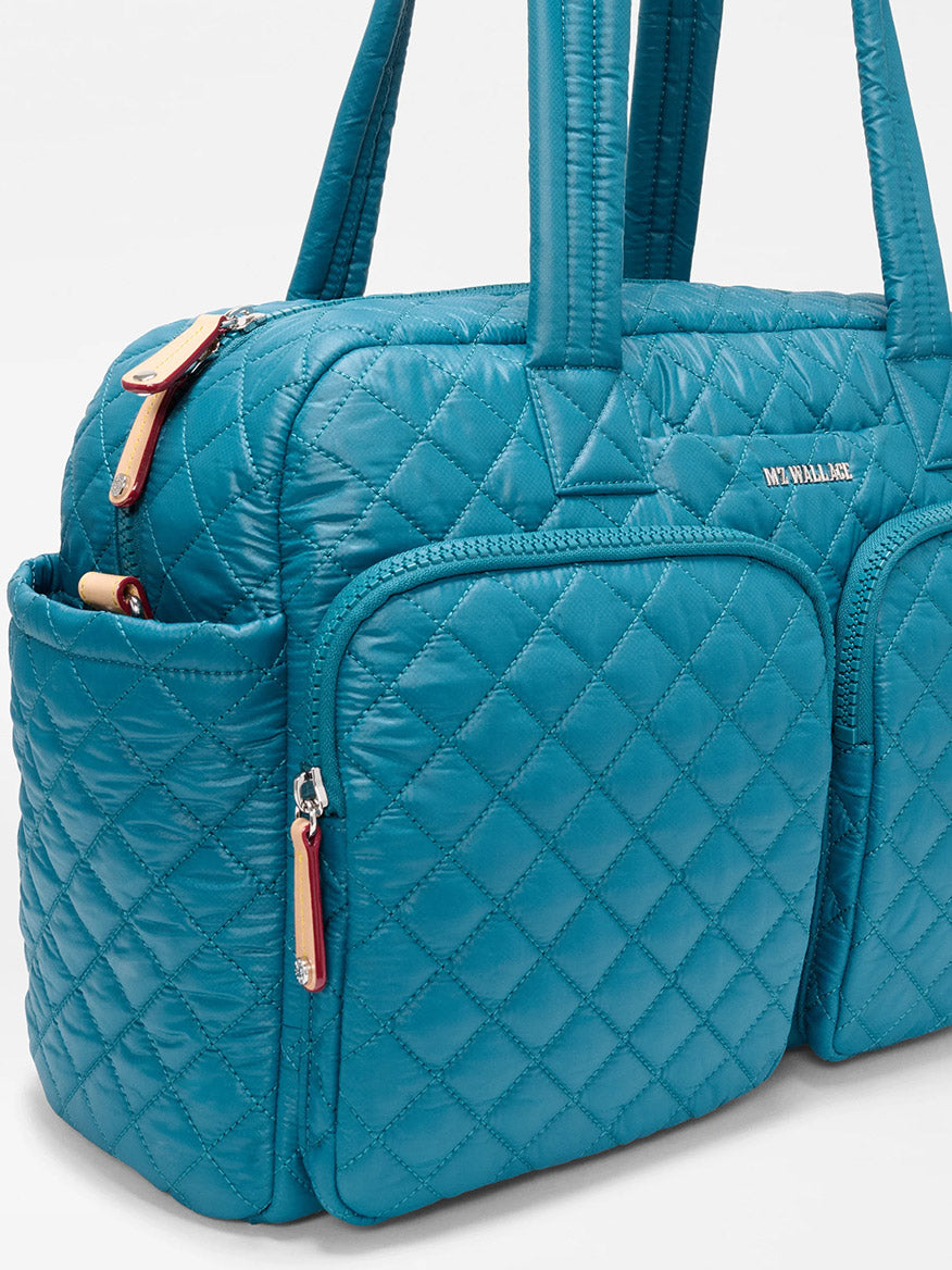 A close-up of the MZ Wallace Nik Duffel in Pacific Oxford shows its blue quilted design, two front zippered pockets, a side pocket, and long padded handles.