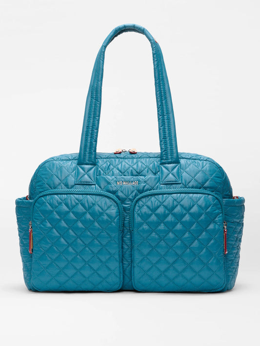 The MZ Wallace Nik Duffel in Pacific Oxford is a teal quilted tote with two front zippered pockets, side pockets, and long handles—ideal as a travel carryall—shown against a plain white background.