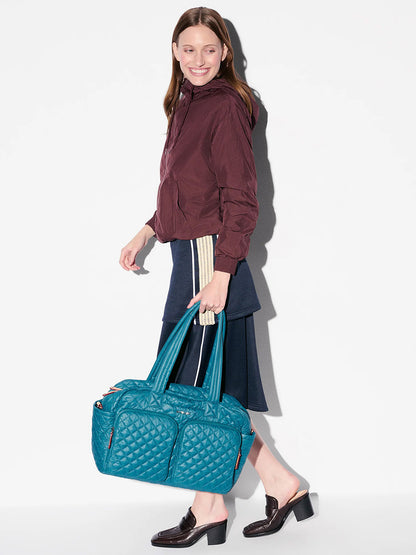 A woman in a maroon jacket and navy skirt smiles while carrying the MZ Wallace Nik Duffel in Pacific Oxford.