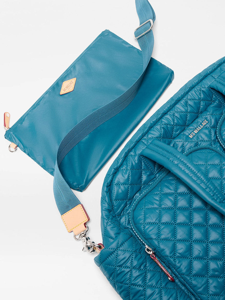 The MZ Wallace Nik Duffel in Pacific Oxford, a quilted blue carryall with a front zipper pocket, sits next to a flat blue pouch with a thick strap and metal clasp, both displayed on a white background.