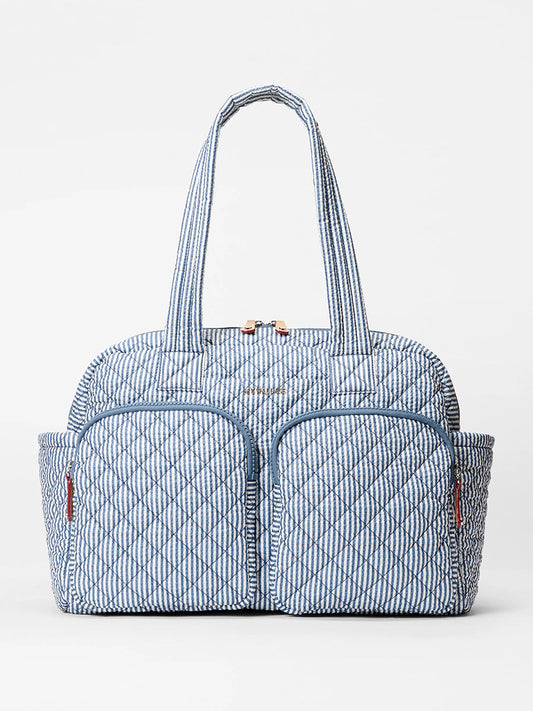 The MZ Wallace Nik Duffel in Seersucker Oxford is a blue and white quilted fabric tote with two front zippered pockets and two side pockets, shown against a plain white background.