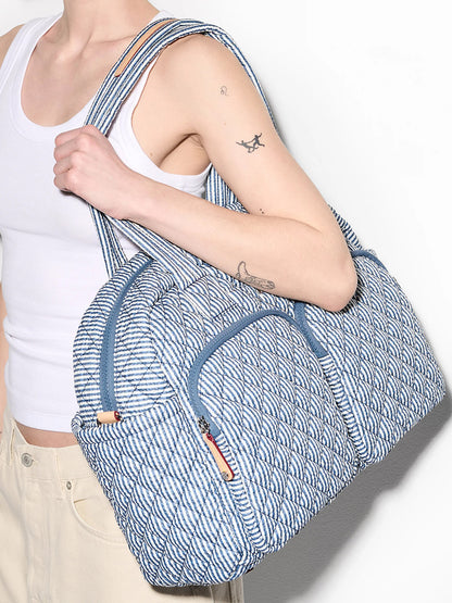 A person in a white tank top and light pants carries the MZ Wallace Nik Duffel in Seersucker Oxford over their shoulder.
