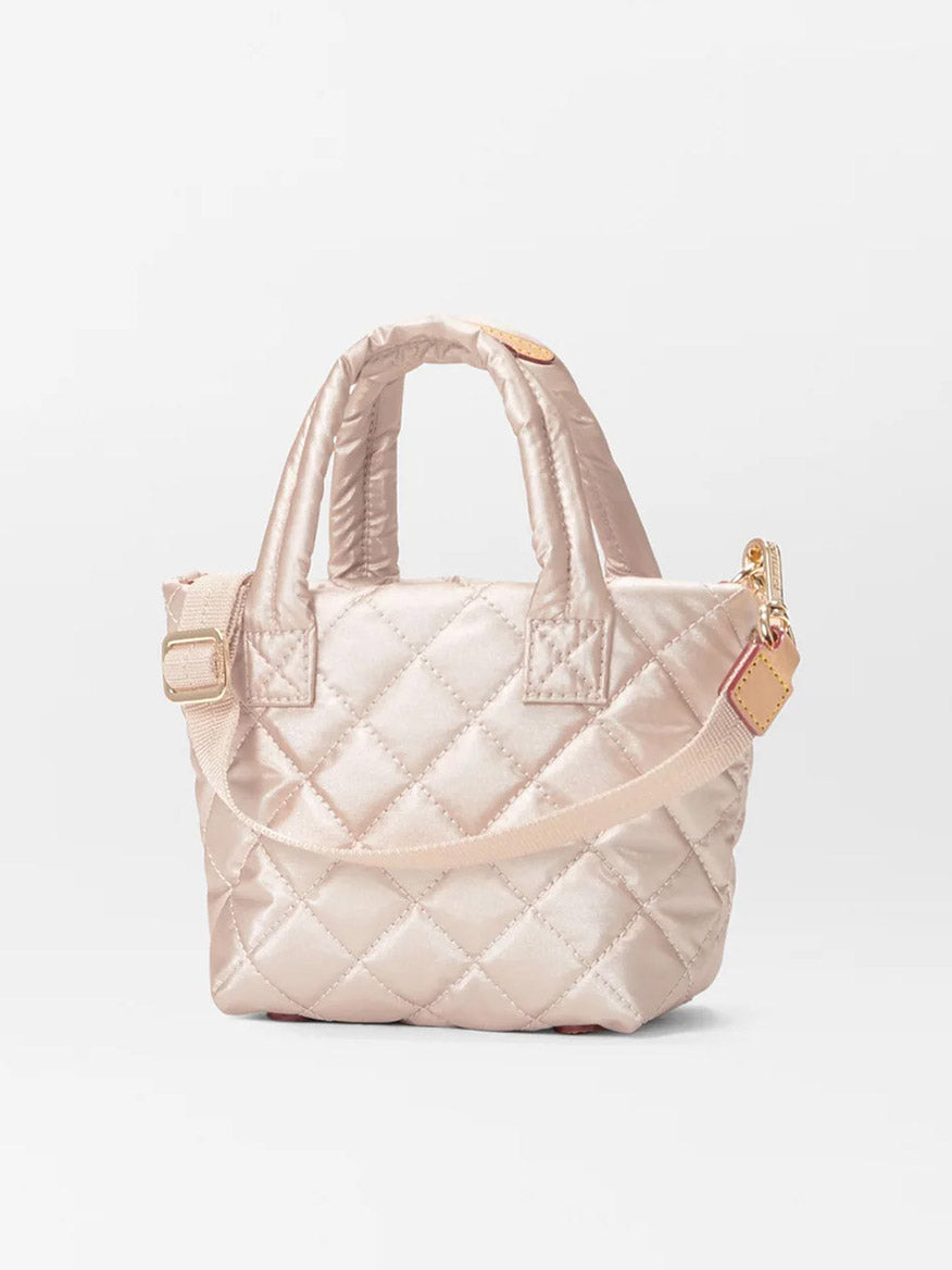 Petite Metro Tote Deluxe in Pale Rose Gold Metallic Oxford by MZ Wallace, a small quilted handbag designed with two handles and an adjustable crossbody strap. It features gold-tone hardware set against a plain white background, crafted from recycled oxford material for an eco-friendly appeal.