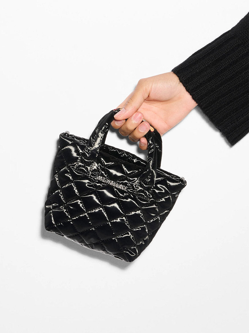 A hand holds the stylish MZ Wallace Petite Metro Tote Deluxe in Black Liquid Oxford, showcasing its quilted design and short handles against a plain background.