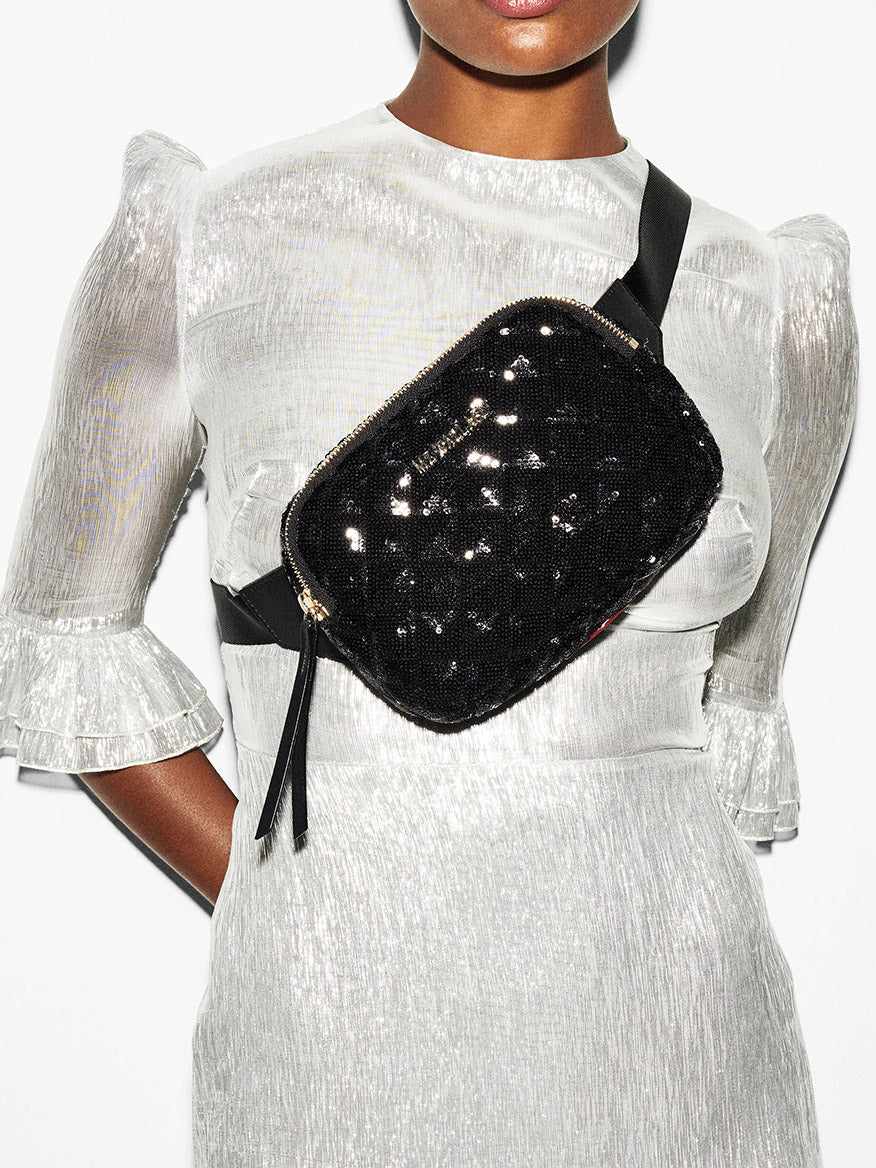 A close-up of a woman wearing a shimmering silver dress and an MZ Wallace Quilted Madison Belt Bag in Black Sequin with Italian leather trim over her shoulder.