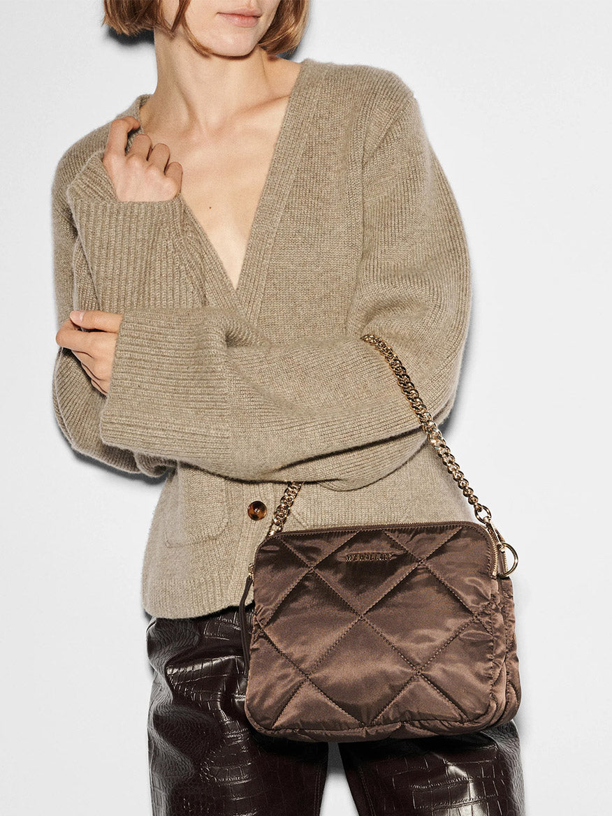 A person in a beige sweater holds the MZ Wallace Quilted Madison Crossbody in Walnut Bedford, highlighting its elegant brown design with a detachable chain strap.