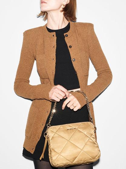 Woman in a brown jacket over a black dress, holding a gold MZ Wallace Quilted Madison Crossbody in Camel Bedford. Only the torso is visible.