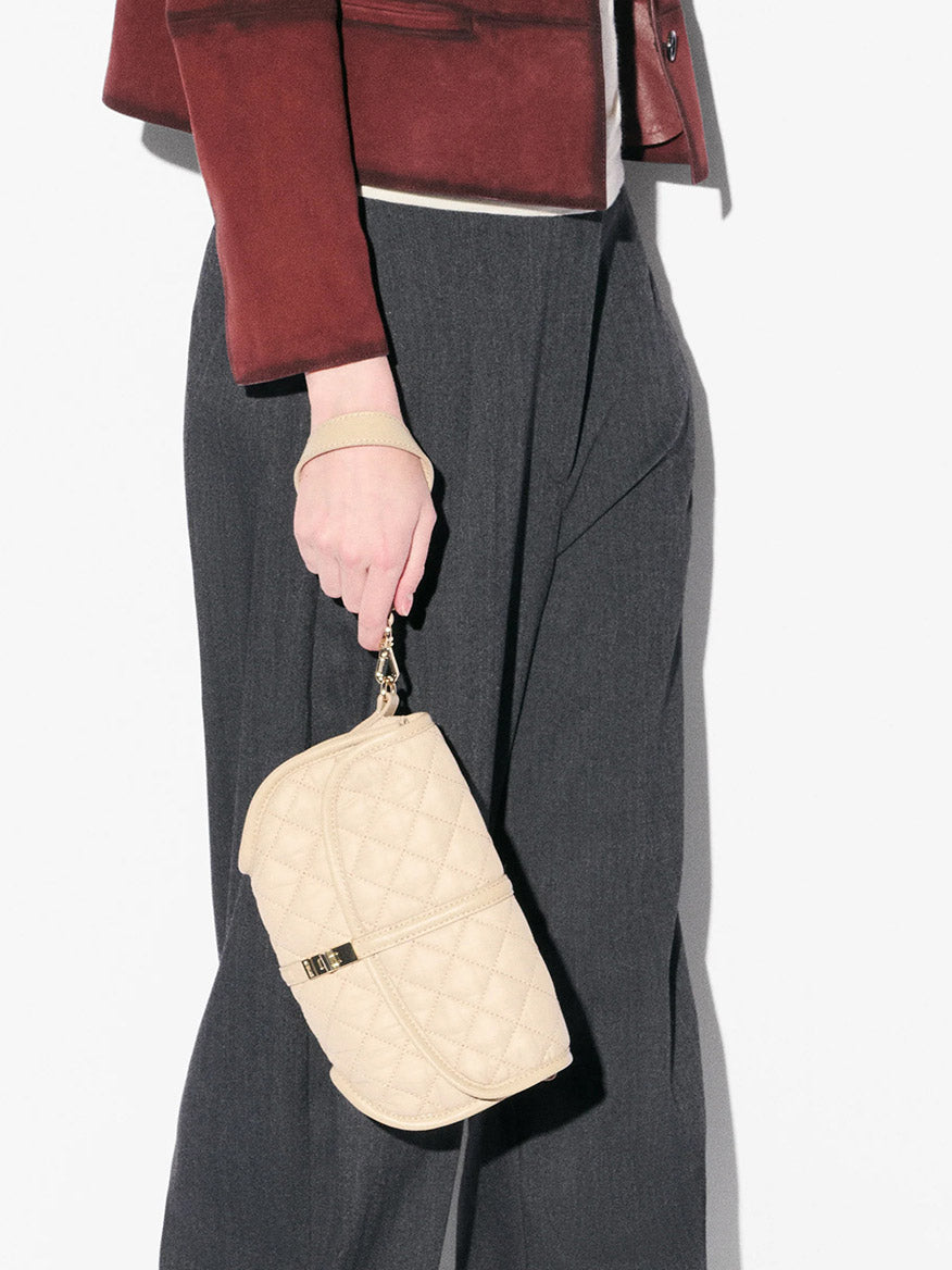 A person in a burgundy jacket and gray pants holds the MZ Wallace Small Astor Convertible Clutch in Buff Oxford, featuring an Italian leather strap and quilted design, against a plain background.