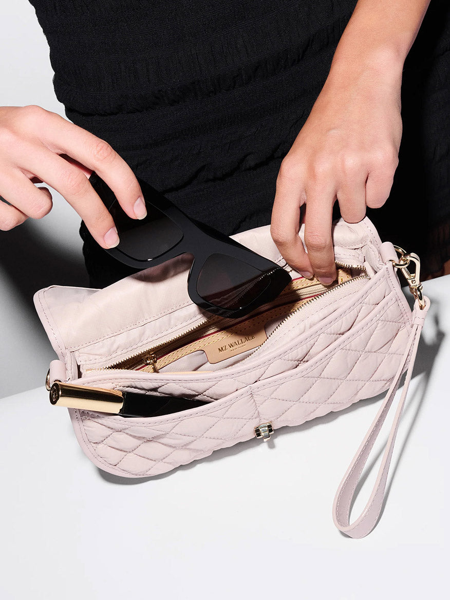 A person places black sunglasses into an MZ Wallace Small Astor Convertible Clutch in City Pink Oxford, which is partially unzipped to reveal its contents.