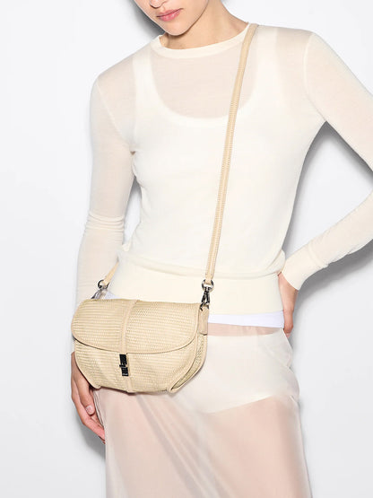 Person wearing a sheer long-sleeve white top, white skirt, and carrying the MZ Wallace Small Astor Convertible Clutch in Buff Mesh—a light beige crossbody with adjustable strap and flap closure.