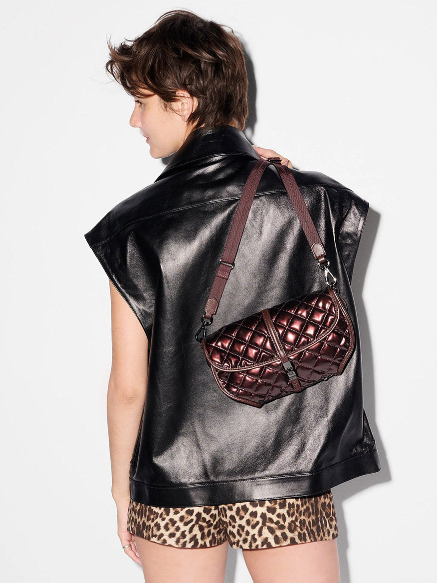 A person wearing a black leather vest and leopard print shorts is shown from the back, carrying the MZ Wallace Small Astor Convertible Clutch in Bordeaux Lacquer Oxford over their shoulder.