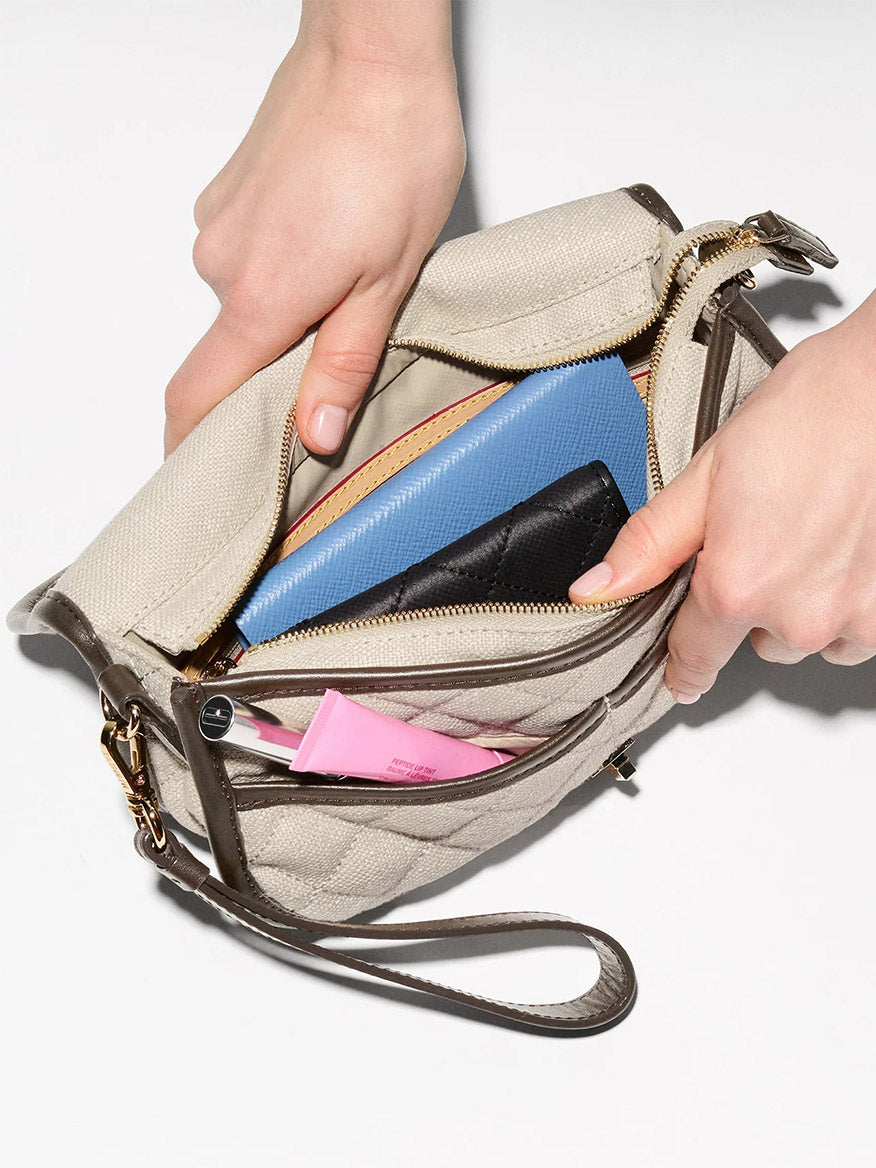 A person opens the MZ Wallace Small Astor Convertible Clutch in sandy linen fabric with Italian leather trim, revealing a blue wallet, a black item, and a pink tube—a stylish summer essential.