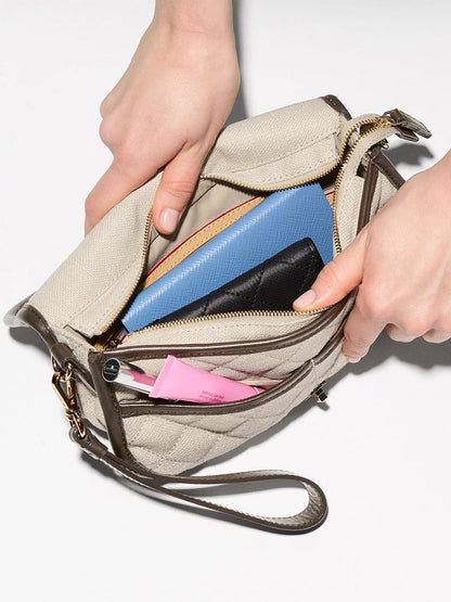 A person opens the MZ Wallace Small Astor Convertible Clutch in sandy linen fabric with Italian leather trim, revealing a blue wallet, a black item, and a pink tube—a stylish summer essential.