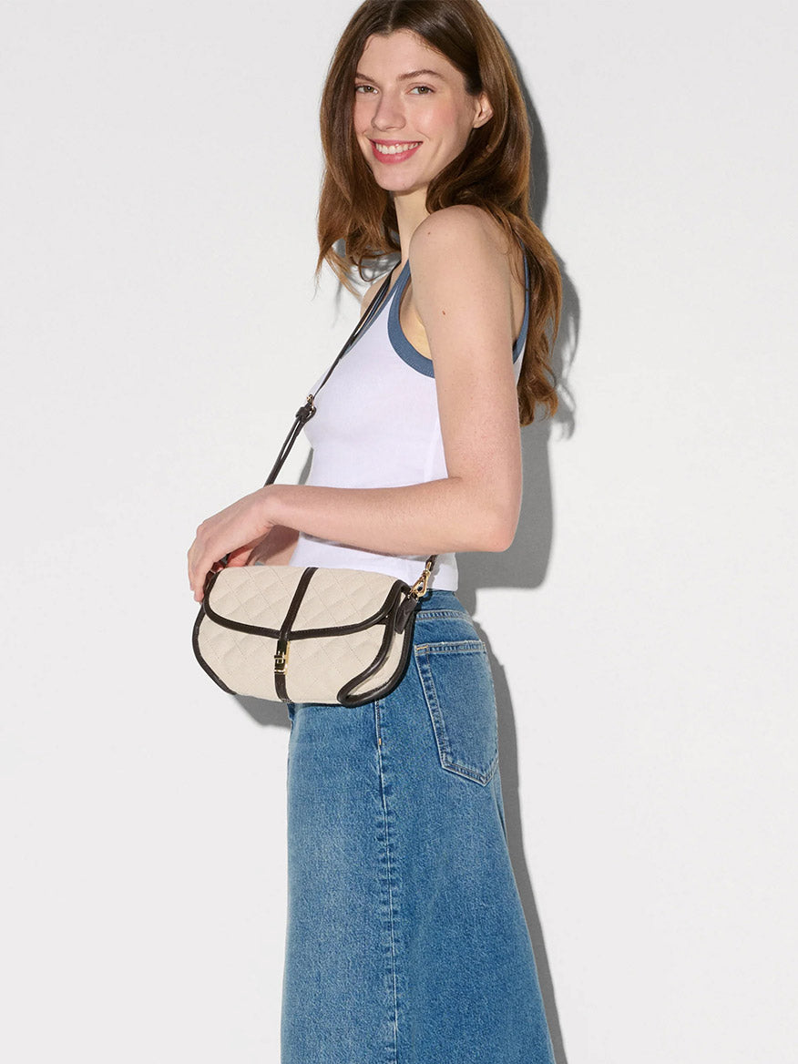 A woman in a white tank top and blue denim skirt holds the MZ Wallace Small Astor Convertible Clutch in Sandy Linen Fabric, featuring beige and black accents with Italian leather trim—a summer classic against a plain light background.