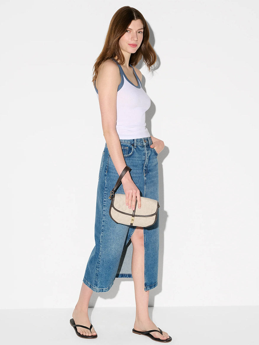 A woman in a white tank top, denim midi skirt, and black flip-flops stands against a plain background, holding the MZ Wallace Small Astor Convertible Clutch in Sandy Linen Fabric—a true summer classic.