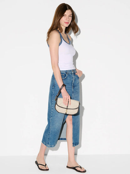 A woman in a white tank top, denim midi skirt, and black flip-flops stands against a plain background, holding the MZ Wallace Small Astor Convertible Clutch in Sandy Linen Fabric—a true summer classic.