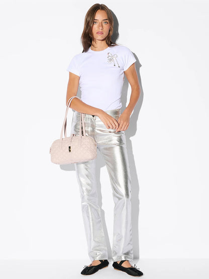 A woman stands against a white background wearing a white t-shirt, metallic silver pants, black flats, and carrying the MZ Wallace Small Astor Satchel in City Pink Oxford.