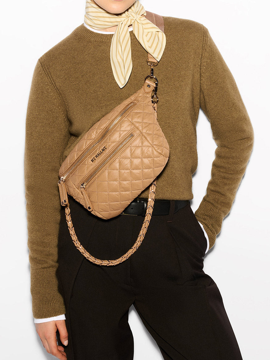 A person wearing a brown sweater, striped scarf, and black pants is carrying an MZ Wallace Small Crosby Sling Bag in Caramel Oxford across their chest.