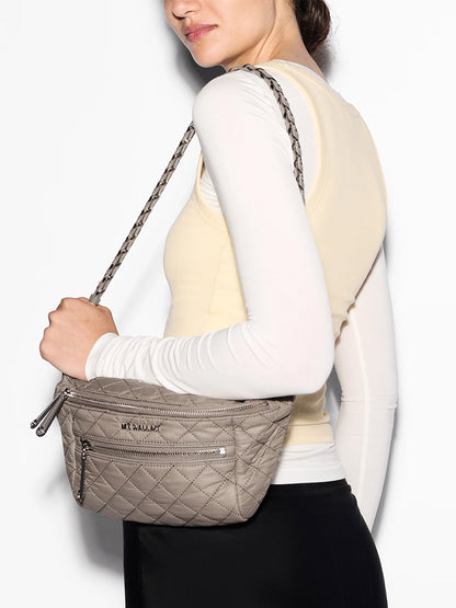 A person wearing a sleeveless beige top over a long-sleeve white shirt carries the MZ Wallace Small Crosby Sling Bag in Morning Dove Oxford, a crossbody bag with quilted fabric and zippered pockets.