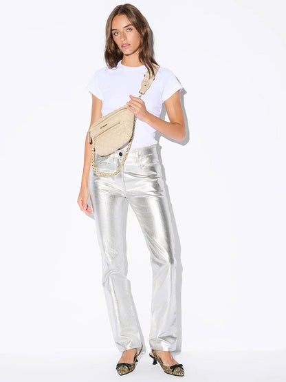 A woman stands against a white background wearing a white t-shirt, shiny silver pants, pointed shoes, and carrying the MZ Wallace Small Crosby Sling Bag in Buff Oxford.
