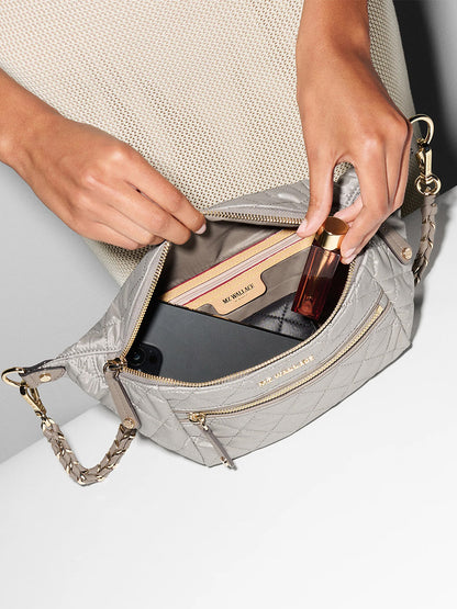 A person opens an MZ Wallace Small Crosby Sling Bag in Sable Pearl Metallic Oxford, revealing a beige wallet, a small bottle, and a smartphone inside.