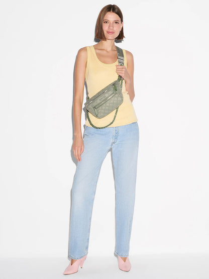 A person stands against a white background wearing a yellow tank top, light blue jeans, pink heels, and carrying the MZ Wallace Small Crosby Sling Bag in Agave Green Oxford with an adjustable strap.