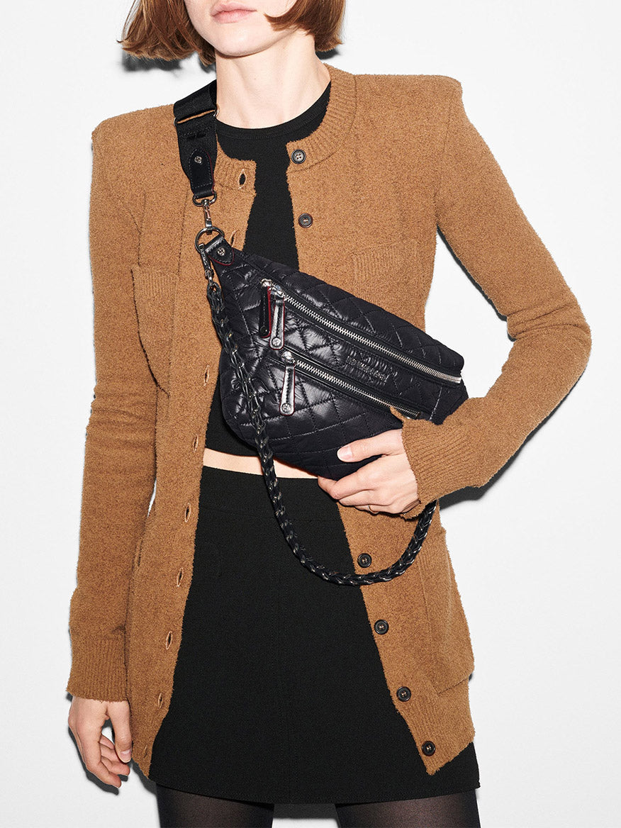 A person wearing a brown cardigan and black skirt holds an MZ Wallace Small Crosby Sling Bag in Black Oxford, featuring exterior pockets and a crossbody strap.