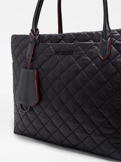 Close-up of the MZ Wallace Small Daily Tote in Black Oxford, a quilted black tote bag crafted from recycled nylon with leather handles, subtle red trim, and an attached tag. The “MZ WALLACE” logo is embossed on the front, blending sophisticated style with everyday functionality.