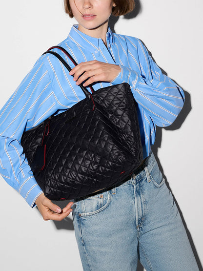 A person wearing a blue striped shirt and light jeans holds the MZ Wallace Small Daily Tote in Black Oxford, crafted from recycled nylon with two handles, against a plain background.