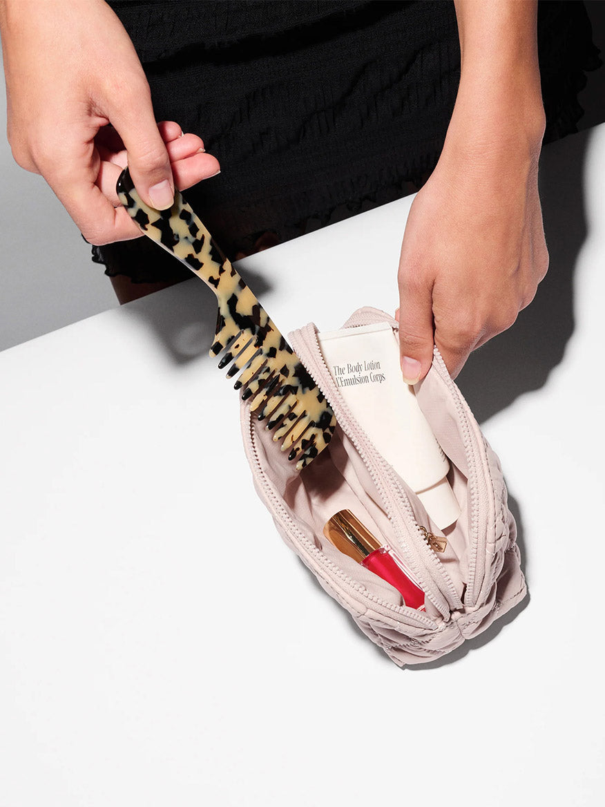 A person puts a tortoiseshell-patterned comb into the MZ Wallace Small Double Zip Cosmetic in City Pink Oxford, a light pink zippered pouch, on a white surface.