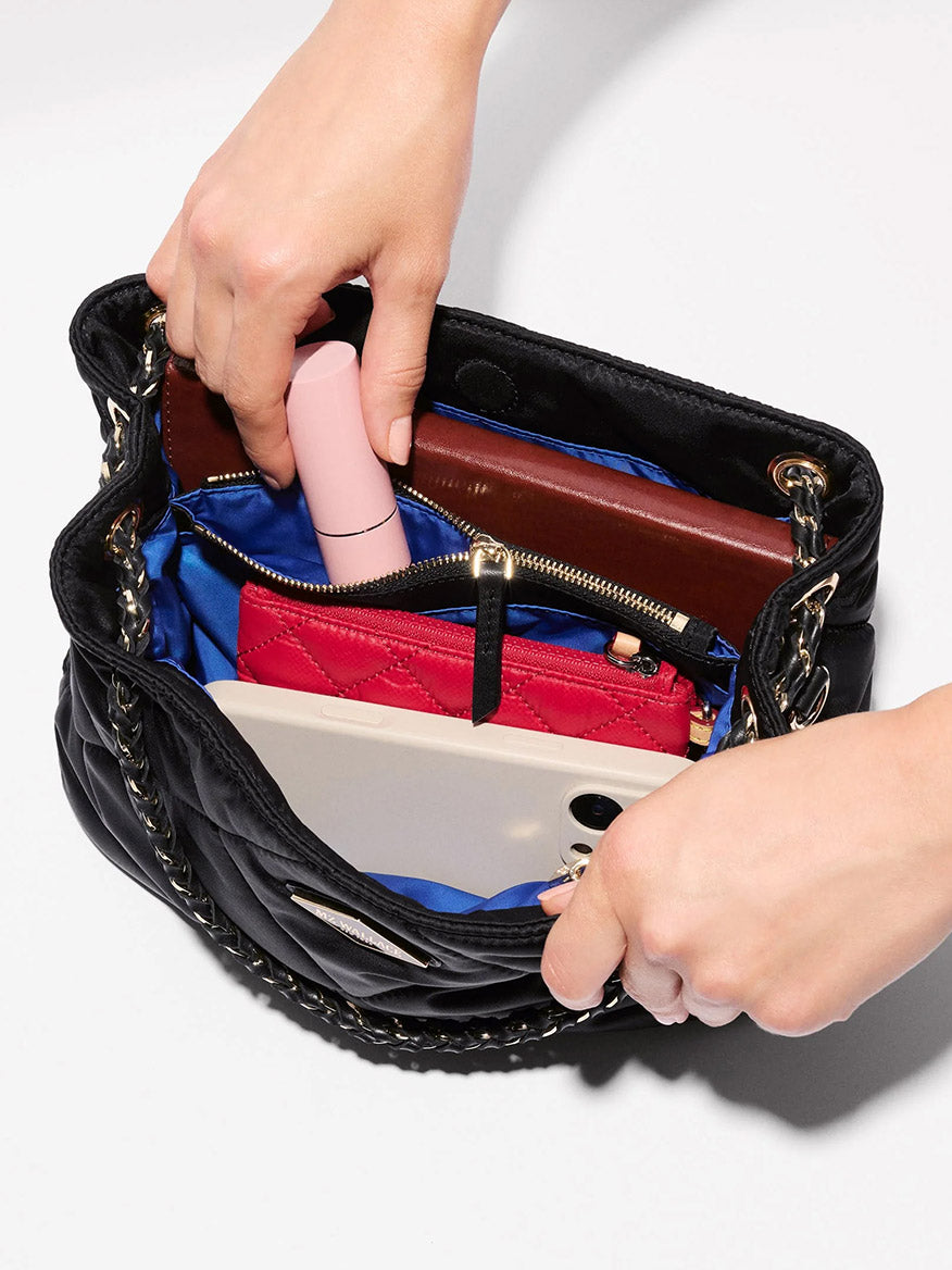 A person opens the MZ Wallace Small Frankie Tote in Black Bedford, a compact handbag crafted from quilted nylon. Inside, a wallet, a pink cylindrical item, a red pouch, and a smartphone are displayed against a white background. The adjustable crossbody strap offers added convenience and versatility to this stylish tote.