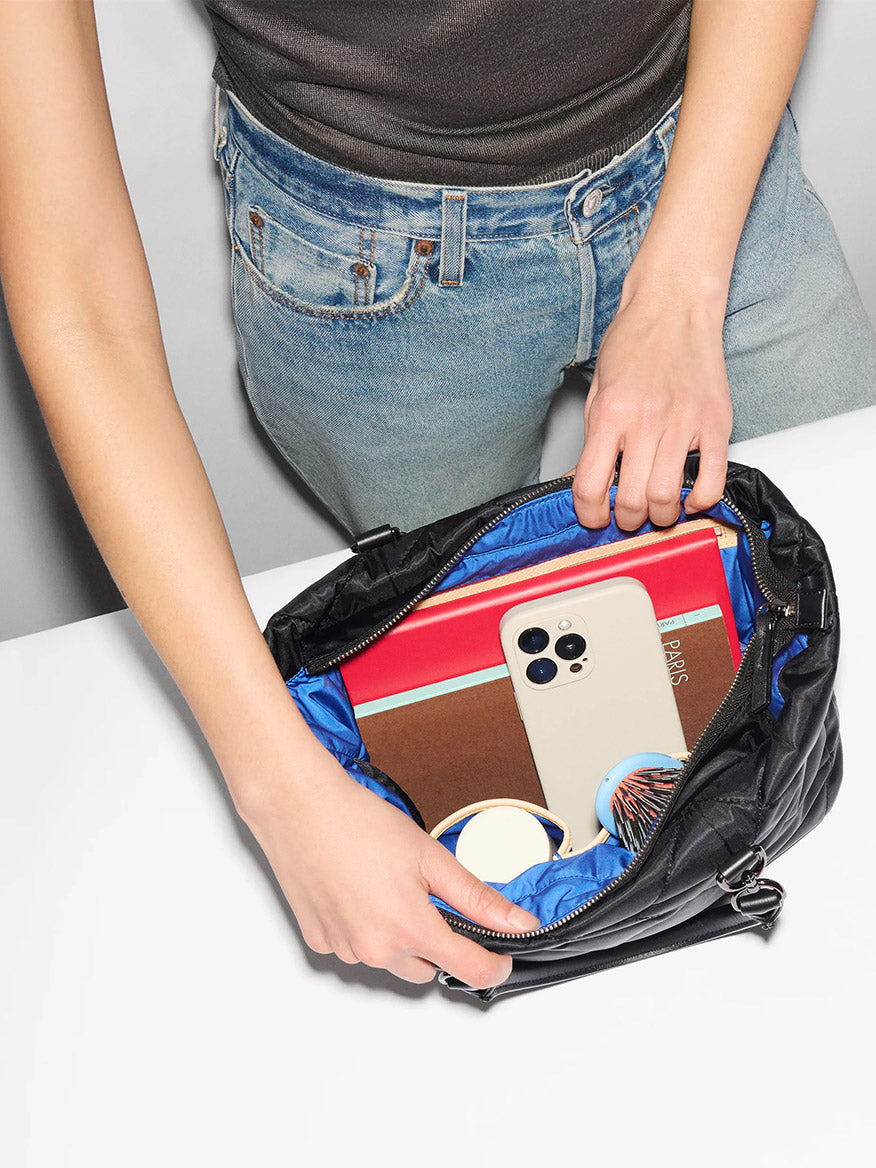 A person holds open the MZ Wallace Small Madison E/W Tote in Black Bedford, revealing a phone, red notebook, brown wallet, striped pouch, and two round containers inside its slim silhouette with Italian leather trim.