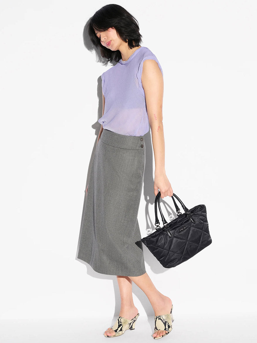 Woman wearing a light purple sleeveless top, gray midi skirt, and snakeskin heels stands against a white background, holding the MZ Wallace Small Madison E/W Tote in Black Bedford with Italian leather trim.