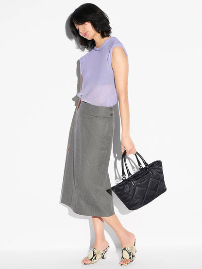 Woman wearing a light purple sleeveless top, gray midi skirt, and snakeskin heels stands against a white background, holding the MZ Wallace Small Madison E/W Tote in Black Bedford with Italian leather trim.
