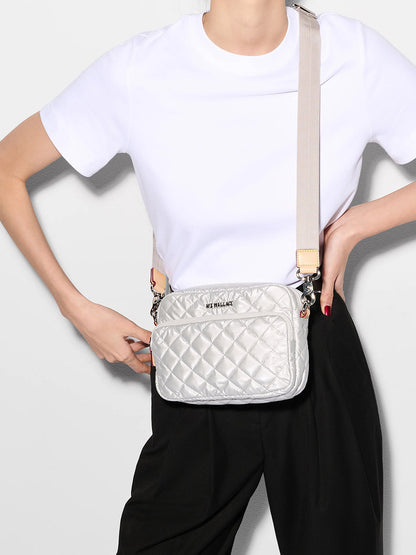 A person in a white t-shirt and black pants models the MZ Wallace Small Metro Camera Bag in Oyster Metallic Oxford, which features quilted silver nylon, leather trim, and a beige strap.