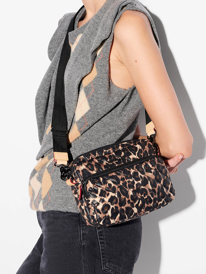 A person in a sleeveless gray argyle sweater and black jeans carries the MZ Wallace Small Metro Camera Bag in Leopard Oxford as a crossbody, featuring a sleek black strap.