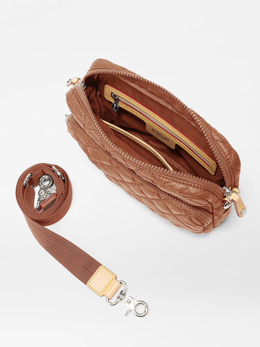 The MZ Wallace Small Metro Camera Bag in Terracotta Oxford is a lightweight, brown quilted nylon crossbody. It features an open zipper revealing a striped lining and interior pockets. The bag includes a detachable adjustable strap with metal clips, offering versatile style for any outing.