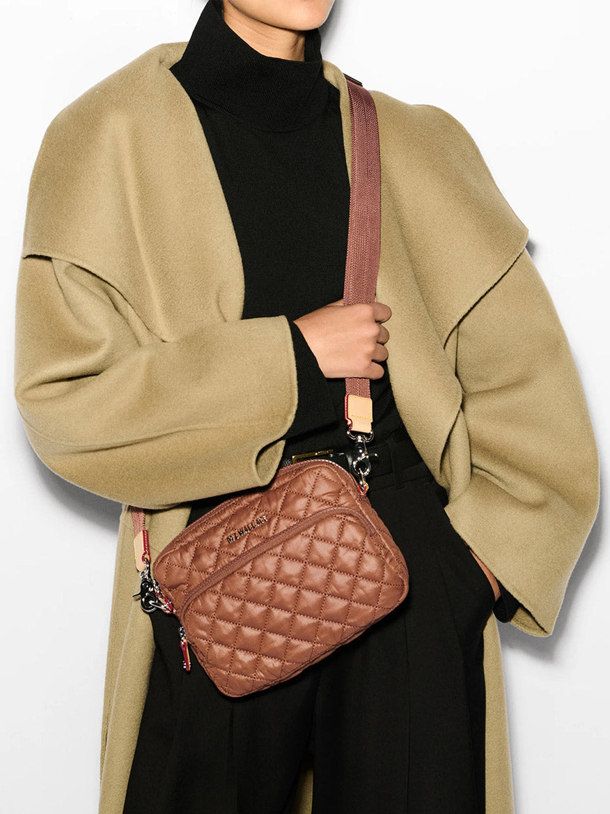 A person in a beige coat and black top carries an MZ Wallace Small Metro Camera Bag in Terracotta Oxford, featuring a quilted nylon design and an adjustable wide strap.