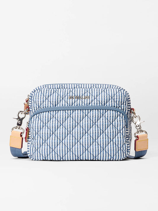 The MZ Wallace Small Metro Camera Bag in Seersucker Oxford features blue and white vertical stripes, tan leather accents, quilted nylon material, and a front zipper pocket, displayed on a plain background.