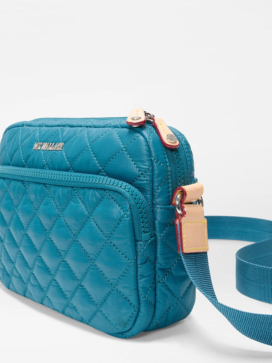 The MZ Wallace Small Metro Camera Bag in Pacific Oxford features teal quilted nylon, a front zip pocket, gold-tone zipper, and an adjustable strap, shown as a compact crossbody against a plain white background.
