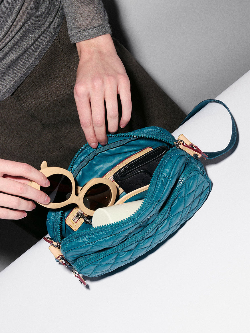 Someone places round beige sunglasses into an open MZ Wallace Small Metro Camera Bag in Pacific Oxford on a white surface, with a black wallet and a light-colored comb inside the bag.