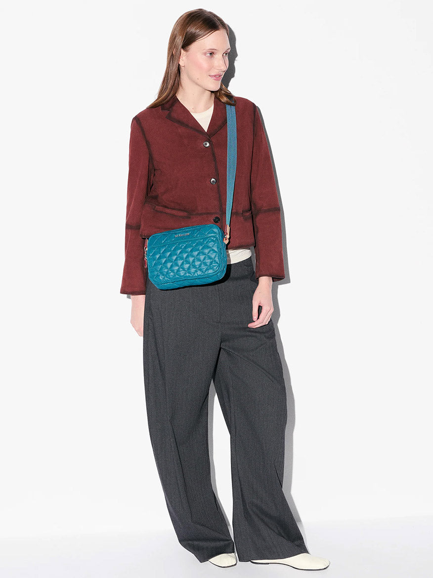 A woman stands against a white background, wearing a maroon jacket, gray wide-leg pants, white shoes, and carrying an MZ Wallace Small Metro Camera Bag in Pacific Oxford.