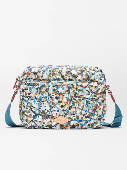 The MZ Wallace Small Metro Camera Bag in Club Paint Oxford features a rectangular nylon design with a blue strap and a multicolored abstract pattern, displayed against a white background.