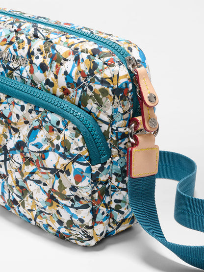 A close-up of the MZ Wallace Small Metro Camera Bag in Club Paint Oxford, featuring a multicolored pattern, nylon material, teal zippers and strap, and a beige leather accent near the strap attachment.
