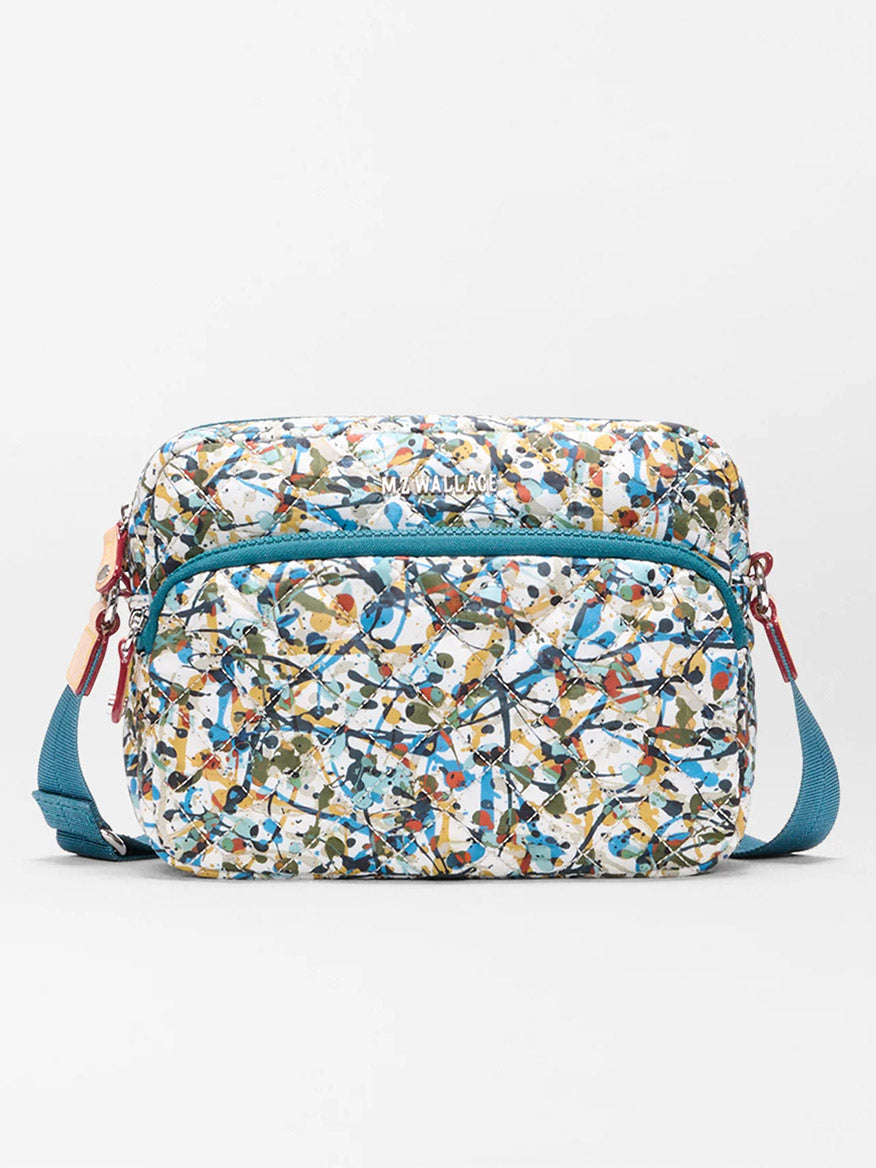 The MZ Wallace Small Metro Camera Bag in Club Paint Oxford features a multicolored abstract pattern, front zip pocket, and teal adjustable strap, displayed against a plain white background.