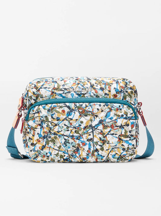 The MZ Wallace Small Metro Camera Bag in Club Paint Oxford features a multicolored abstract pattern, front zip pocket, and teal adjustable strap, displayed against a plain white background.