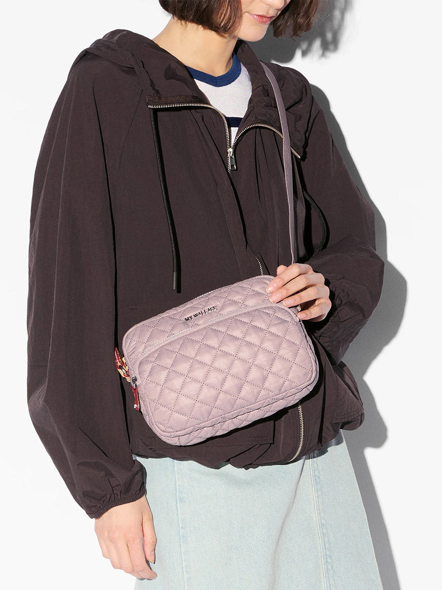 A person wearing a black zip-up jacket and light blue jeans holds the MZ Wallace Small Metro Camera Bag in Quail Oxford.