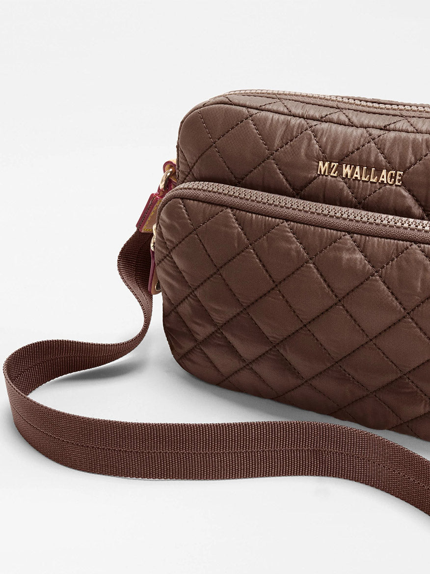 Shown against a plain background is the MZ Wallace Small Metro Camera Bag in Walnut Oxford—a brown quilted recycled nylon bag featuring gold “MZ WALLACE” lettering, a front zipper pocket, and an adjustable strap for hands-free carry.